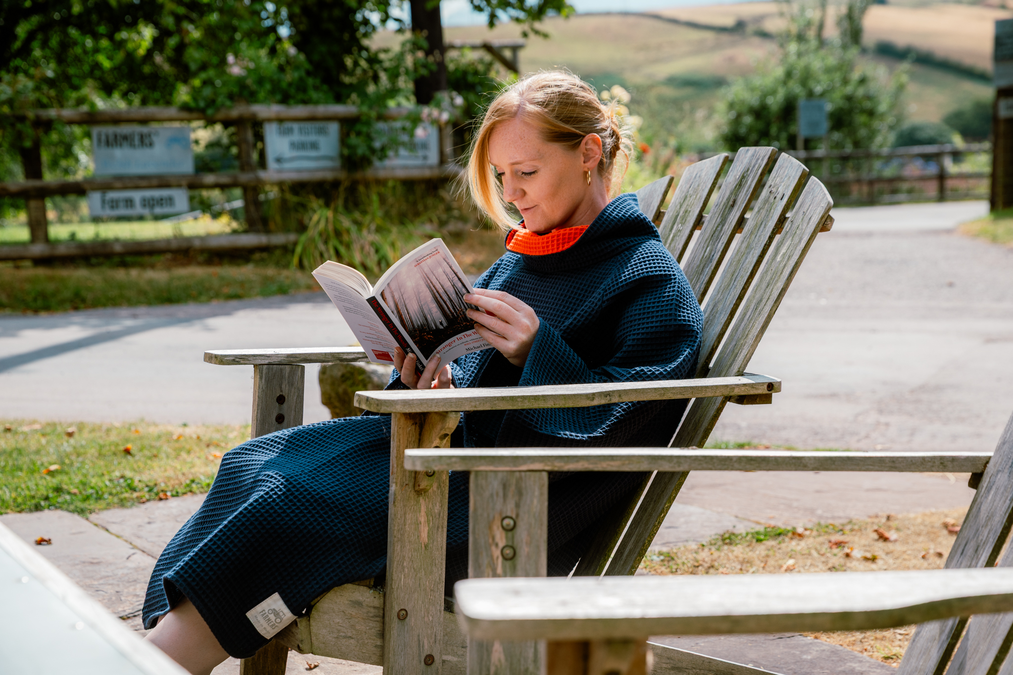 Woman sat in a garden chair wearing a navy FARMERS' Waffle robe and reading a book